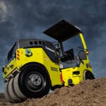 A yellow and gray Vacker Neuson compactor is shown working on a dirt mound under a dark, cloudy sky. The perspective is low and dramatic, emphasizing the compactor's size and the rugged construction machinery terrain.