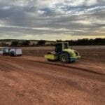 431478954_1443467396248738_4670304030907067173_n - Conplant Pty Ltd Yellow road roller compacting red dirt on a large construction site, with two trailers in the background under a cloudy sunset sky.