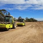 8350abbd-91ee-44fa-bec2-a7759a8cc2eb - Conplant Pty Ltd Two yellow Complant construction vehicles are diligently working on leveling a dirt road under the partly cloudy WA sky. The road is surrounded by trees, with a white vehicle sporting a Garli logo parked on the right side of the image, ensuring the job gets done efficiently.