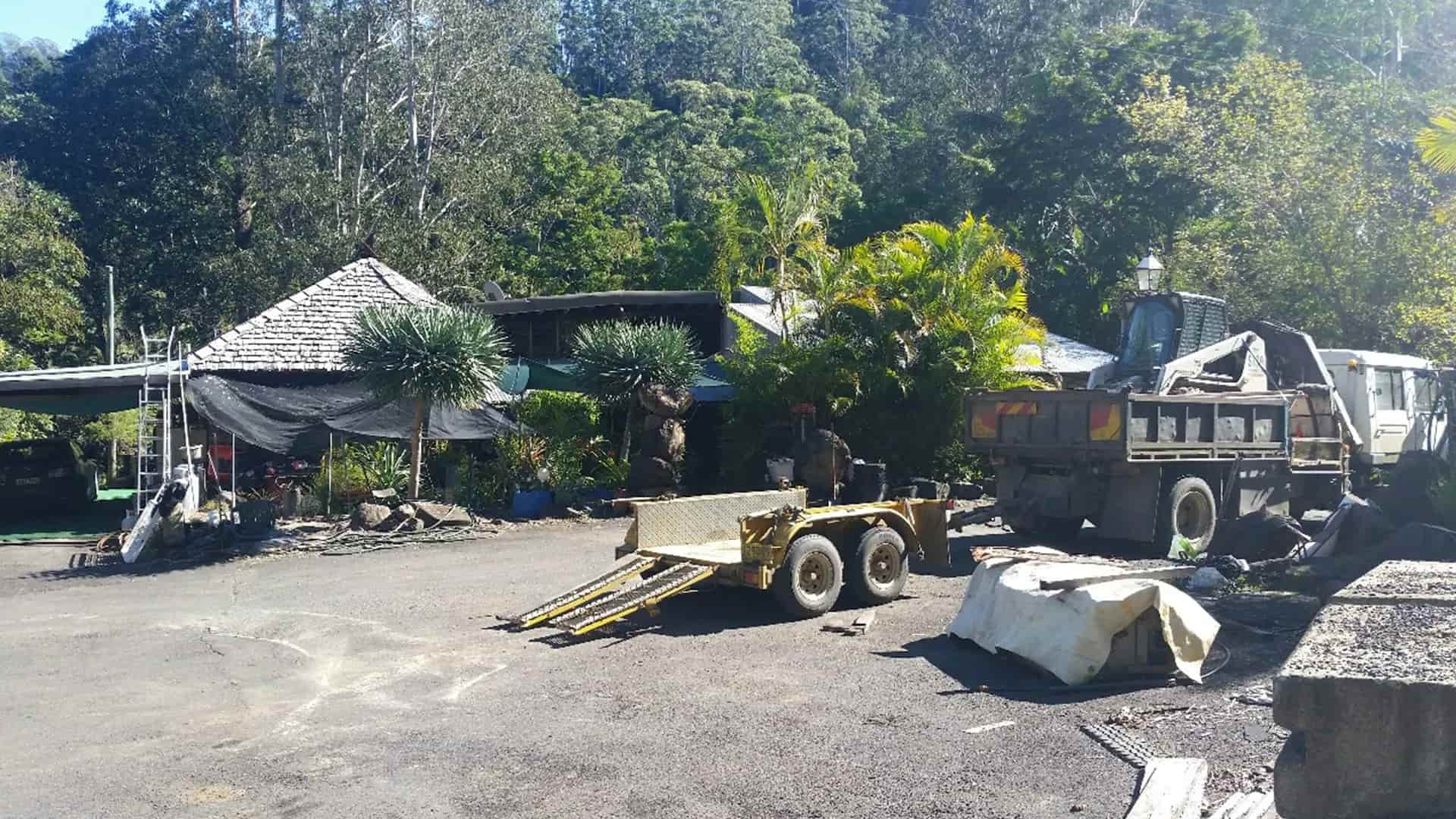 A rural property with a parked truck and trailer in the foreground evokes a scene straight from a Brian Perkins story. Lush green trees and plants surround the area, while a house with slanted roofs peeks from behind the shade. It's a picturesque sunny day.