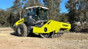 A large, yellow and gray road roller parked on a dirt and gravel surface, surrounded by trees, serves as a guide to effective soil compaction. The machine features a prominent front drum for compacting soil and asphalt, with the word "Compliant" visible on the side.