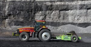 A red and yellow tractor labeled "BH01" is pulling a green Broons Impact Roller in a mining site. The background features layers of exposed rock and a large wall of excavated earth. The ground is covered in dark, loose material typical of mining environments.