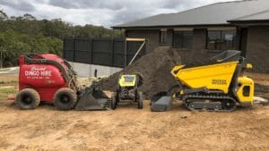 A lineup of construction equipment on a dirt lot in front of a dark brick building includes a red utility machine and a yellow compact track loader. Brady Faulkner oversees the operation, with a mound of soil in the background, framed by trees and overcast skies.