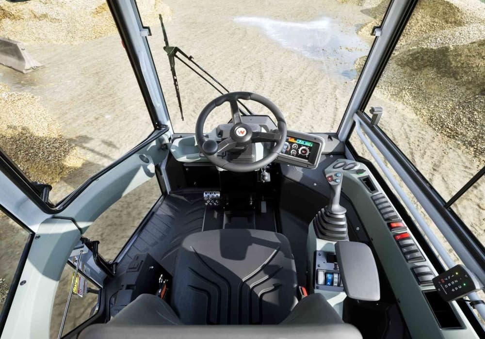 View from above of a modern construction vehicle cab, showing a seat, steering wheel, various levers, control panels, digital display, and windows overlooking a gravel-covered ground outside.