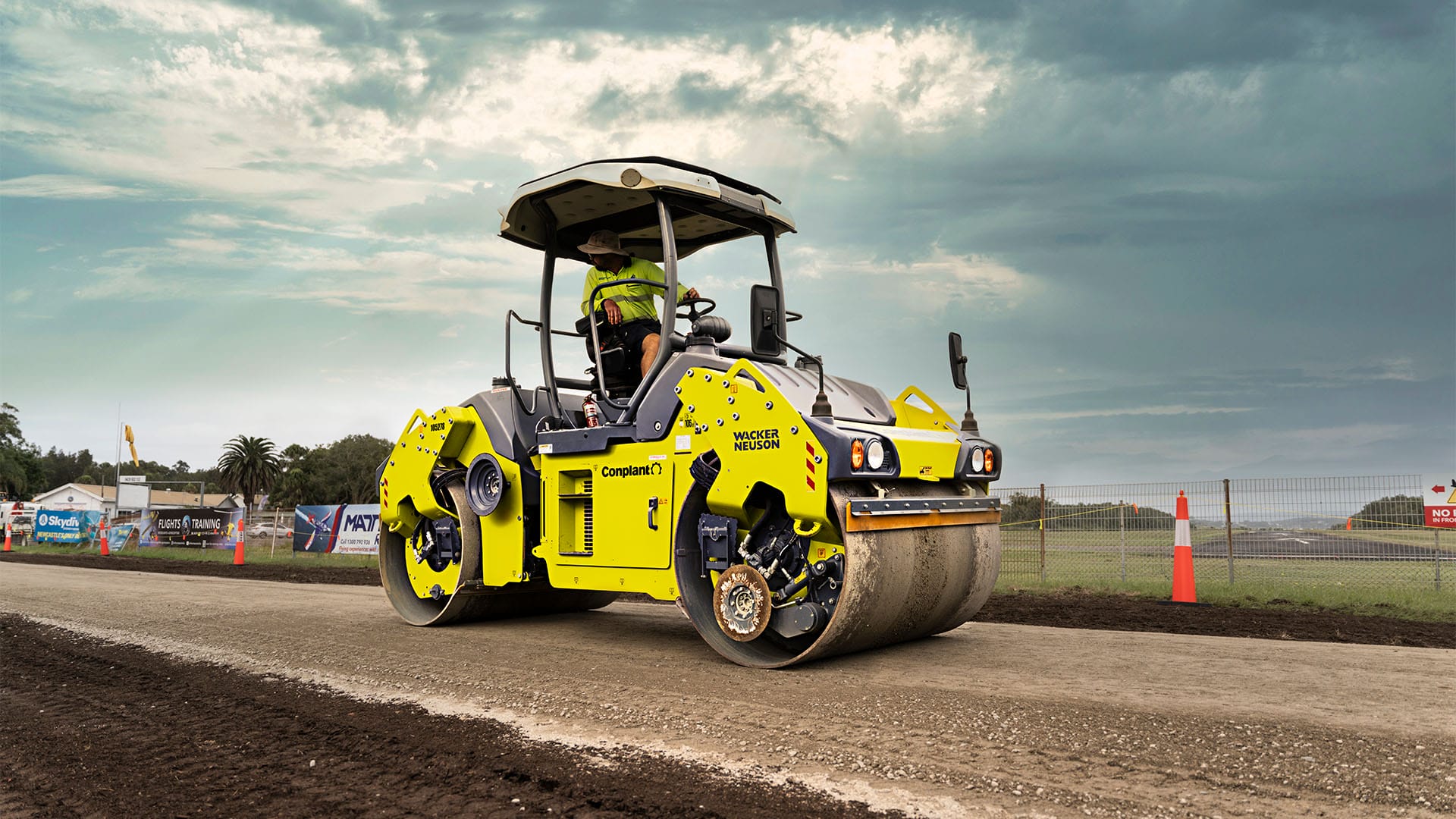 A yellow road roller ensures ideal soil compaction on a dirt road under a cloudy sky.