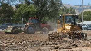 A red tractor pulls a green trailer with tires, while a yellow bulldozer, vital for construction estimation, pushes debris at a bustling site. Trees and hills form the backdrop under a clear sky.