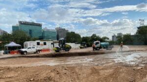 At the Cross River Rail construction site, workers operate machinery on a dirt surface. A water truck sprays the ground as a roller compacts the soil. Portable buildings and city skyscrapers stand in the background beneath a partly cloudy sky.