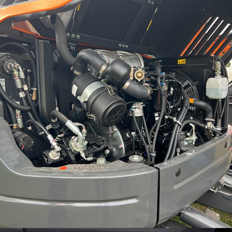 An image showing the exposed engine compartment of a Develon DX80R heavy-duty vehicle. The engine is surrounded by various pipes, hoses, and components, with a focus on the intricate network of mechanical parts inside the machinery's frame.