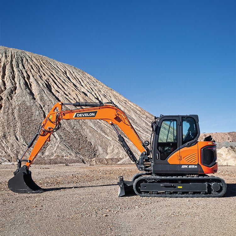 An orange Develon DX89R-7 excavator is parked on a gravel surface with a large, rugged dirt mound in the background under a clear blue sky. The excavator’s arm and bucket are extended forward.