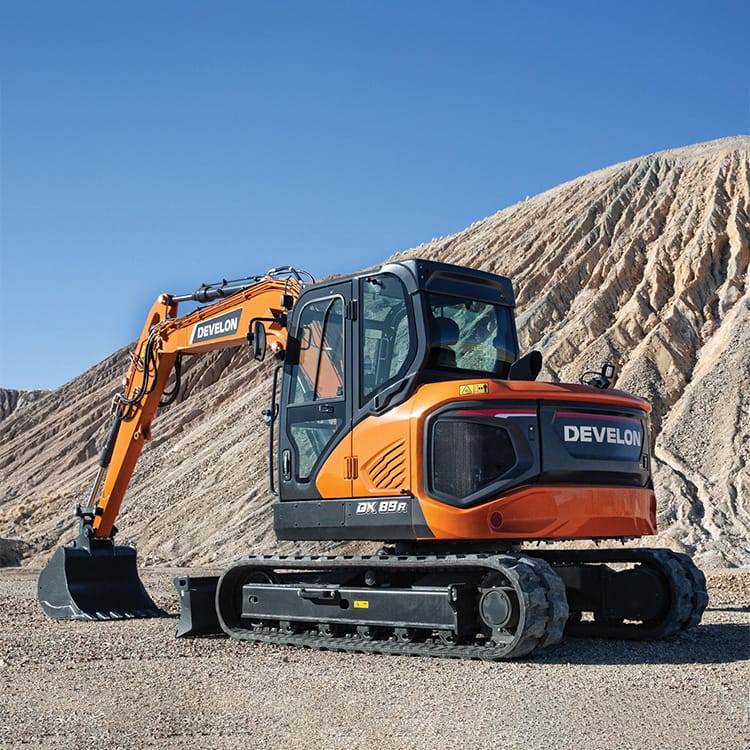 A bright orange Develon DX89R-7 construction excavator sits at a rocky construction site with a large, rugged mound in the background under a clear blue sky. The machine features a tracked base and a hydraulic arm with a digging bucket attachment.