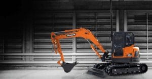 An orange Develon Mini Excavator with a bucket attachment is parked indoors against a wall with horizontal metal panels and railings, as seen at a national dealer.
