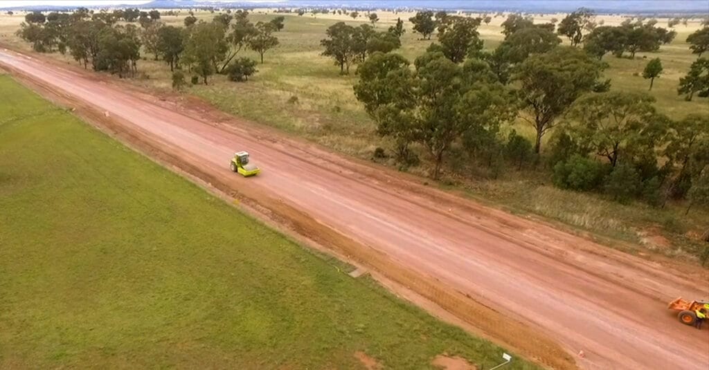 An aerial view of a rural area reveals a dirt road under construction, with soil being moved by two construction vehicles—one in the foreground and the other in the background. The road, showing early corrugations, winds through grassy fields and clusters of trees under an overcast sky.