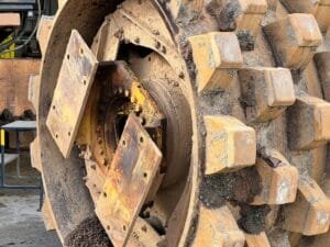 Close-up view of a large, weathered gear wheel with a rugged, textured surface. The blocky teeth, caked in dirt and rust, are clear signs you need new equipment. Mounted vertically, the gear is part of heavy industrial machinery.