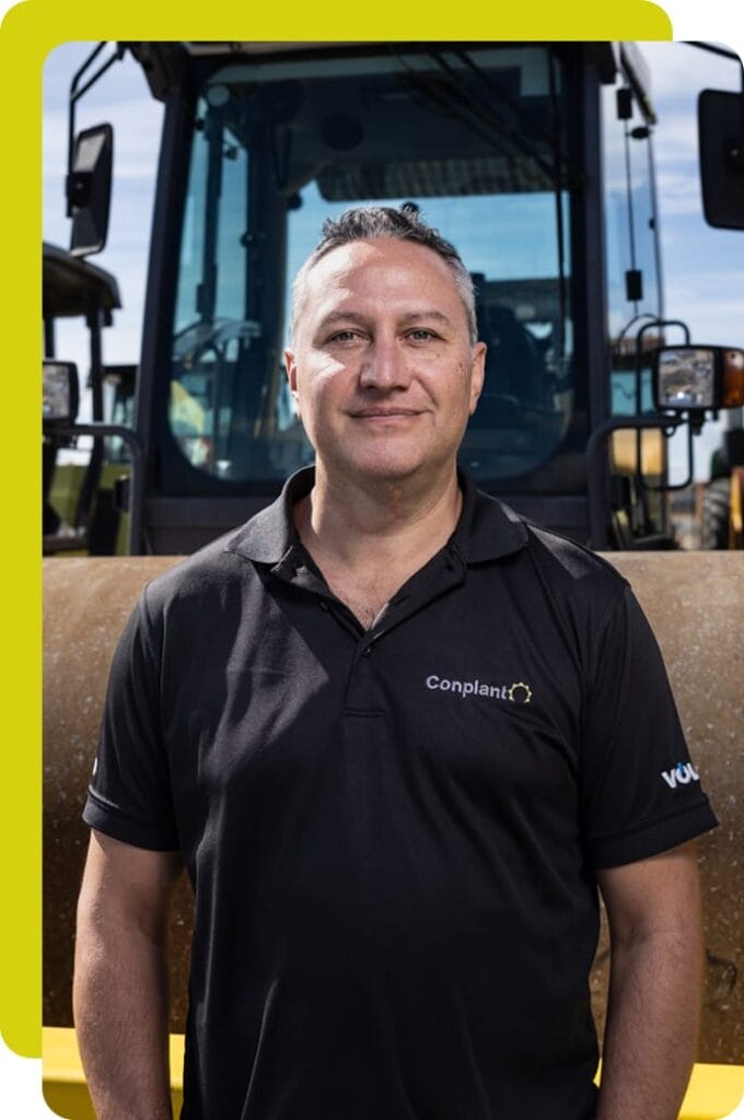 Liam Brosnan stands in front of heavy machinery, wearing a black polo shirt with the Conplant logo. Behind him, parts of construction equipment are silhouetted against a clear blue sky.