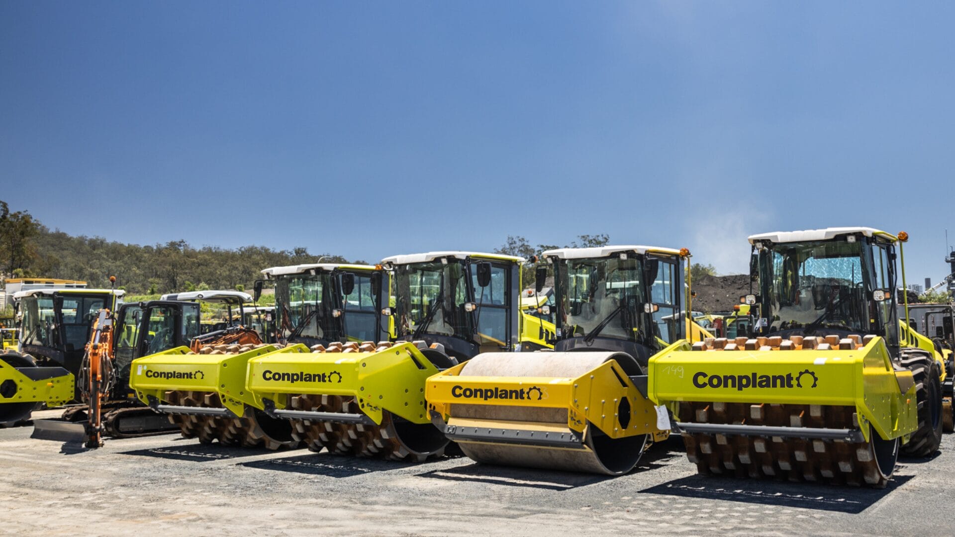 A row of bright yellow construction vehicles, including steamrollers and other heavy equipment, are neatly lined up in a heavy equipment storage area on a dusty lot under a clear blue sky. "Conplant" is visible on the vehicles, ensuring site safety with trees in the background.