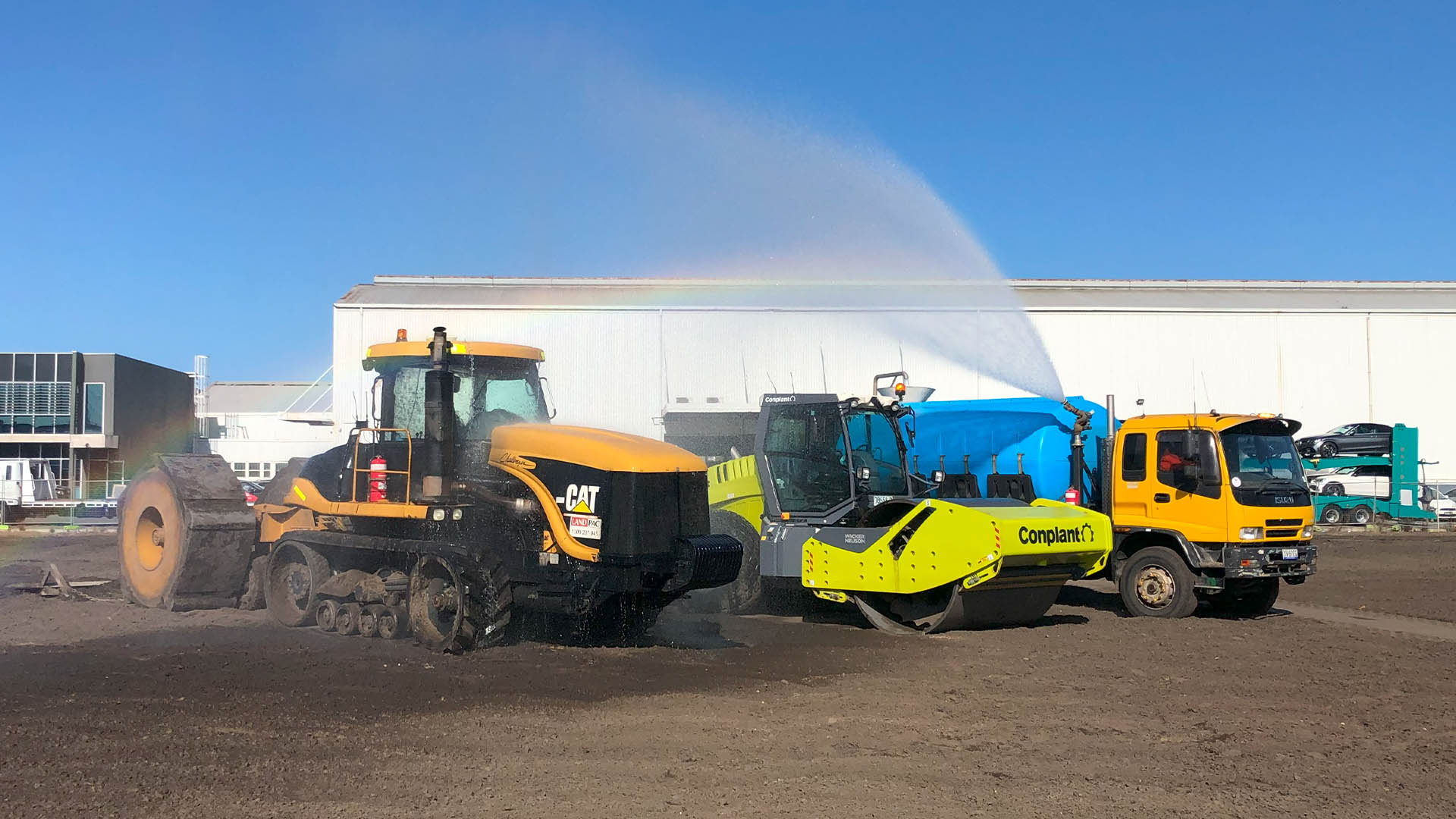 Amidst a bustling construction project, three vehicles operate: a yellow Caterpillar earth mover, a green Conplant roller tackling vibratory roller compaction, and a yellow water truck spraying water to create a small rainbow. A white industrial building stands against the blue sky backdrop.