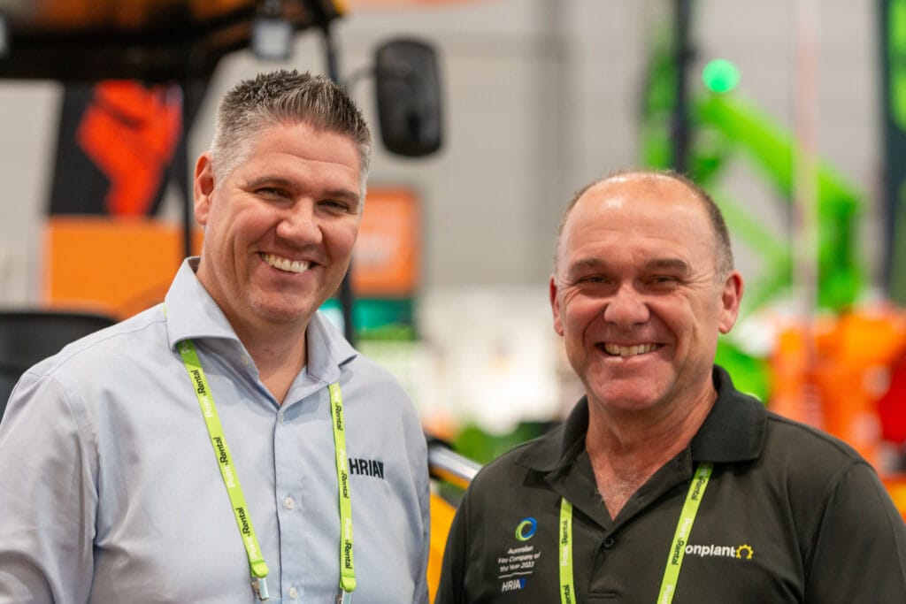 Two men smiling at an indoor event, both wearing collared shirts with company logos and lanyards around their necks. The background is blurred with green and orange equipment visible.