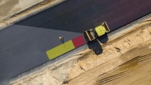 An aerial view showcases a construction vehicle on a freshly paved road. Bordered by sand and dirt, the scene highlights two color patches—green and red—suggesting measurement or coverage. The vehicle might be equipped with an intelligent integrated collision avoidance system for enhanced safety.