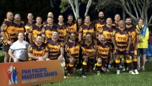 A rugby team in maroon and yellow jerseys poses outdoors, proudly standing and kneeling behind a sign for the Pan Pacific Masters Games on the Gold Coast, Australia. Among them is Jack Alldridge, with trees and grass painting a perfect backdrop.