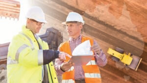 Two construction workers in high-visibility jackets and helmets are examining a tablet and some documents on a construction site. In the background, heavy machinery, a dump truck, and the outline of a map highlight the region's development. One worker points to the tablet screen.