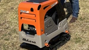 A close-up of a Makasa MVH-308 walk-behind plate compactor on grass showcases the difference in its performance for light and heavy compaction. The machine features an orange, black, and gray color scheme, with part of a person's leg in blue jeans and brown shoes visible in the background.