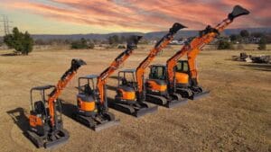 Four orange mini excavators with buckets raised, parked in a row on a vast dirt field, bask under the vibrant rise of a pink and orange sunset. Trees and power lines sketch the background, setting an iconic scene reminiscent of Australia's rugged landscapes.