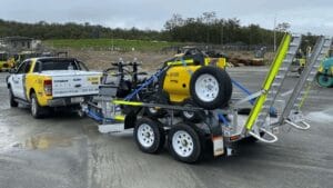 A white utility vehicle is towing a trailer loaded with mini loaders and construction equipment, including a yellow compressor. The trailer has multiple axles and safety ramps. In the background, a construction site brims with machinery and trees.