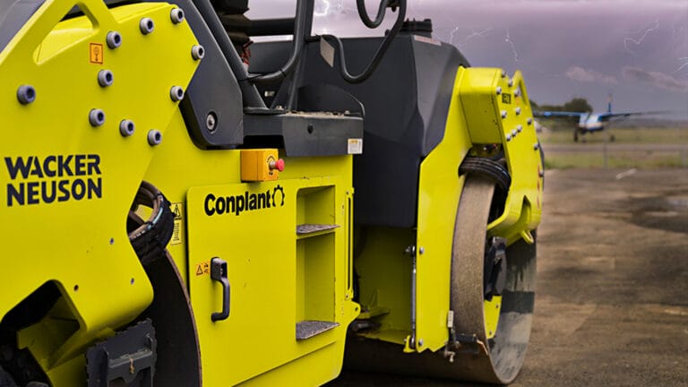 Close-up of a bright yellow construction vehicle, specifically a steamroller, with visible brand names "Wacker Neuson" and "Conplant" on its side. The background shows an airfield with an airplane in the distance, under a cloudy sky with subtle hints of lightning—a vital piece in road maintenance.
