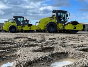 Two vibrant yellow soil compactors parked on a textured dirt surface, with the focus on rugged potholes in the foreground. A partly cloudy sky forms the backdrop, enhancing the industrial setting.