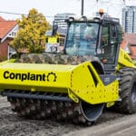 A yellow Conplant road roller, a heavy construction machine equipped with a spiked drum for soil compaction, is smoothing a dirt road in a residential area. Houses and trees can be seen in the background as the roller works efficiently.