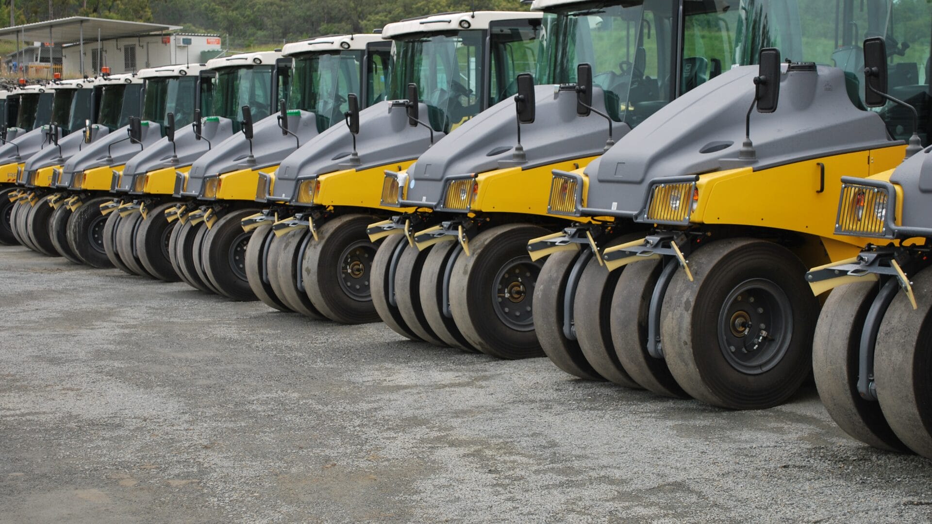 A row of yellow and gray road rollers, including models for pneumatic roller compaction, are parked on a gravel lot, lined up closely next to each other and facing the same direction.
