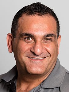 A man with short dark hair and brown eyes, reminiscent of Guy Tome, is smiling at the camera. He is wearing a grey collared shirt and has a light complexion. The background is plain and light-colored.