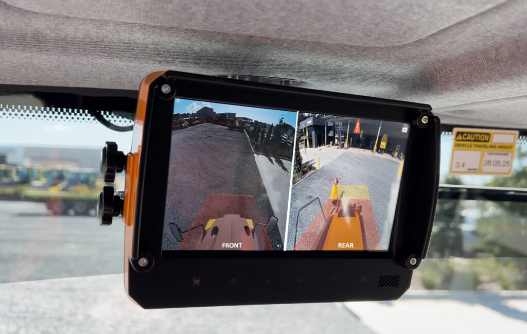 A monitor inside a vehicle displays split-screen front and rear Camera Warning System views of an outdoor area with paved ground, some buildings, traffic cones, and vehicles visible in the distance.