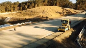 Remote Unmanned 4 - Conplant Pty Ltd A yellow road roller with ROPS2 compacts a newly paved road near a construction site with trees.