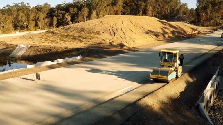 A yellow road roller with ROPS2 compacts a newly paved road near a construction site with trees.