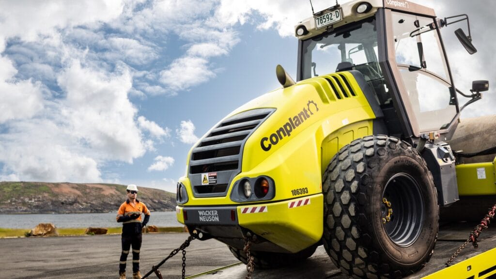 A worker stands by the water near a large compaction roller on a sunny day.