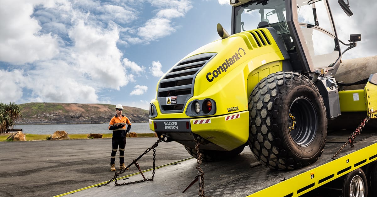 A worker in high-visibility clothing stands near a large yellow construction vehicle marked "Conplant" on a flatbed truck, likely for equipment servicing. The scene is outdoors with some clouds in the sky and a distant mountainous landscape.