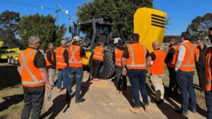 A group of people wearing orange safety vests gathers around a yellow piece of construction equipment with an open hood. They are outdoors on a sunny day, surrounded by trees and a crane, ensuring both safety and precision under the blue sky.