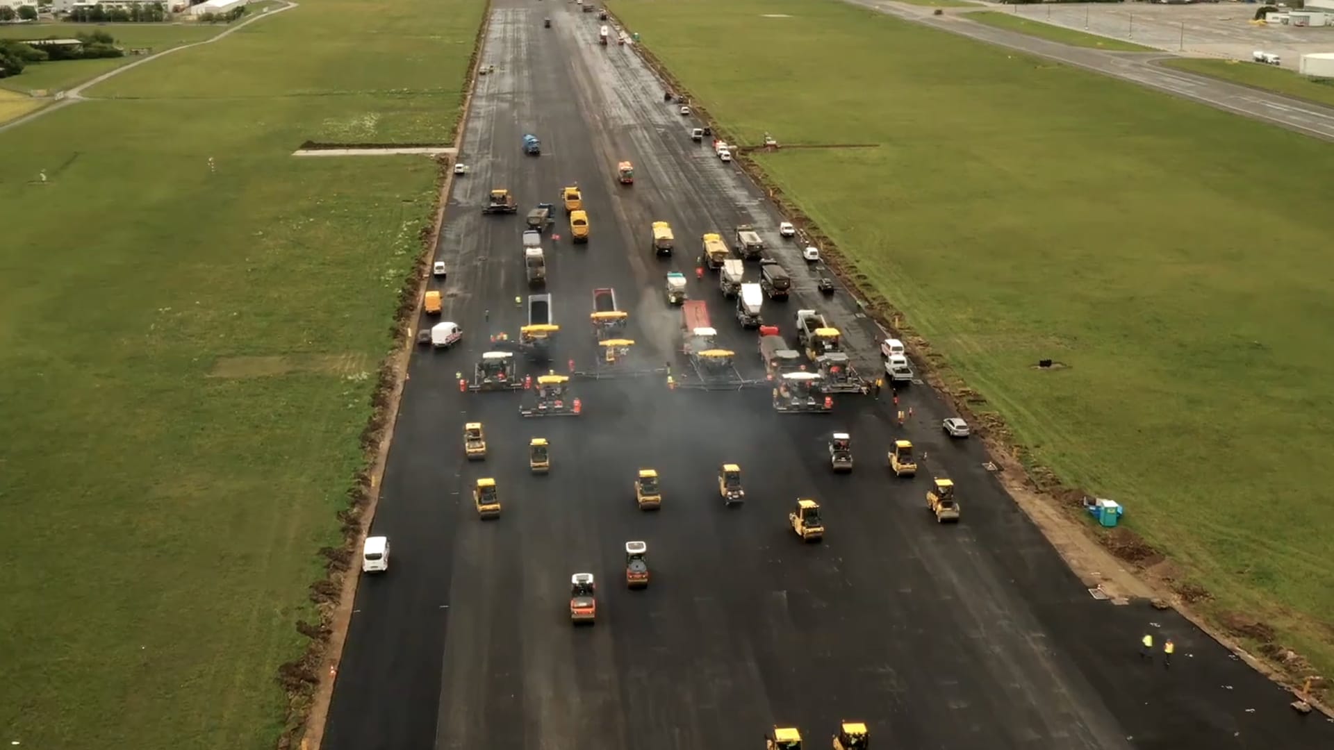 An aerial view captures a runway overhaul at Salzburg Airport, showcasing a large group of construction vehicles working on the wide road surrounded by grassy fields. There's a mix of trucks, rollers, and other machinery, some emitting smoke as they diligently work on the asphalt surface.