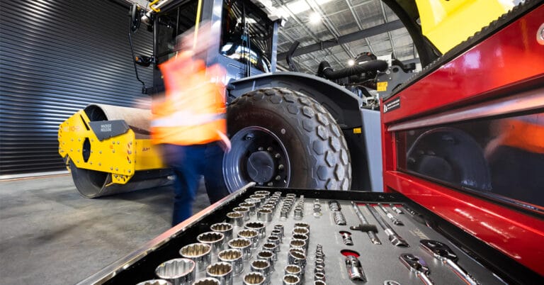 A person in an orange safety vest moves quickly near a large industrial vehicle inside a well-lit garage. In the foreground, an open toolbox displaying various servicing tools, including sockets and wrenches, hints at ongoing equipment maintenance. The background features machinery and a coiled yellow cable.