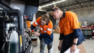 Two workers in high-visibility orange and blue work shirts are examining machinery inside an industrial workshop. One is adjusting parts on the equipment as part of routine equipment maintenance, while the other observes attentively, possibly acting as a guide. The workshop features tools and equipment in the background.