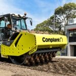 A large yellow compactor machine with the word "Conplant" on it is smoothing a dirt path, ensuring optimal soil compaction. Trees and a modern building are visible in the background under a clear sky.