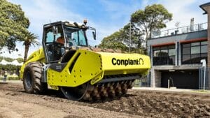 A large yellow compactor machine with the word "Conplant" on it is smoothing a dirt path, ensuring optimal soil compaction. Trees and a modern building are visible in the background under a clear sky.