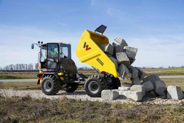 A yellow Wacker Neuson DV125 Dual View Dumper unloads large gray rocks onto a pile outdoors on a gravel surface, with grass and trees in the background under a blue sky.