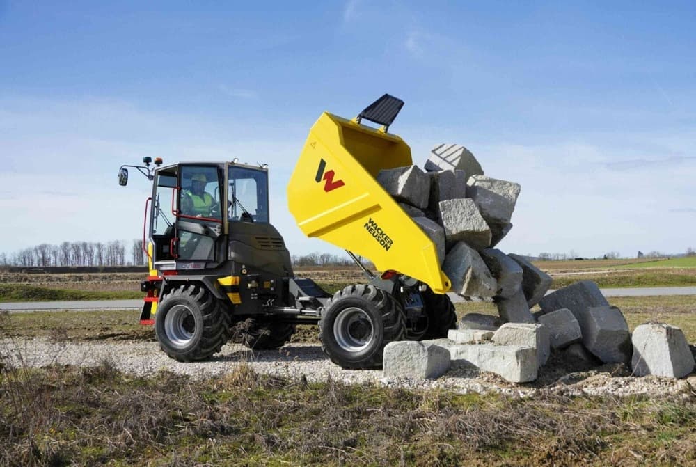 A yellow Wacker Neuson DV125 Dual View Dumper unloads large gray rocks onto a pile outdoors on a gravel surface, with grass and trees in the background under a blue sky.