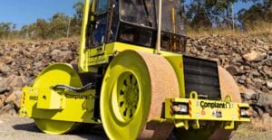 A bright yellow steamroller with large cylindrical wheels stands on a gravel surface, its smooth finish gleaming. Against a rocky backdrop with green foliage and a chain-link fence, this three-point roller is marked with the word "Conplant.