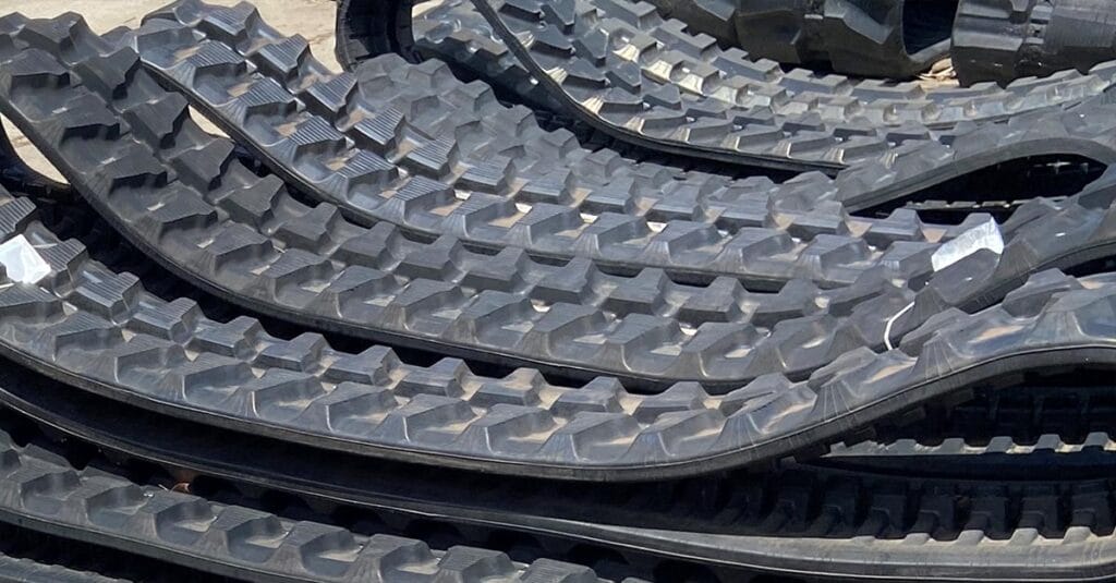 Stacks of large, black rubber tracks with a rugged, treaded surface are piled up in an outdoor setting, likely intended for construction or heavy machinery. The photo captures the texture and intricate patterns of these TrackPads Australia products perfectly.