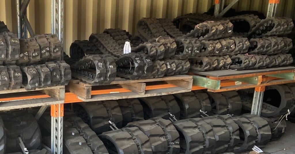 Stacks of industrial rubber tracks labeled with white markings sit on metal shelves in a cluttered warehouse. The arrangement showcases various sizes of tracks, organized and stored in an orderly manner, much like the inventory system at TrackPads Australia during my interview tour.