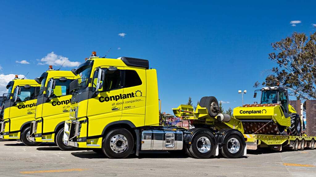 Three bright yellow Conplant trucks and a bulldozer parked outdoors under a blue sky.