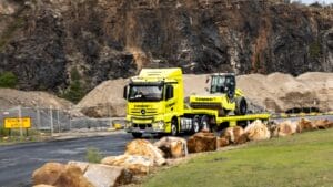 A yellow Conplant truck handles heavy equipment transport past rocks and sand at a quarry.